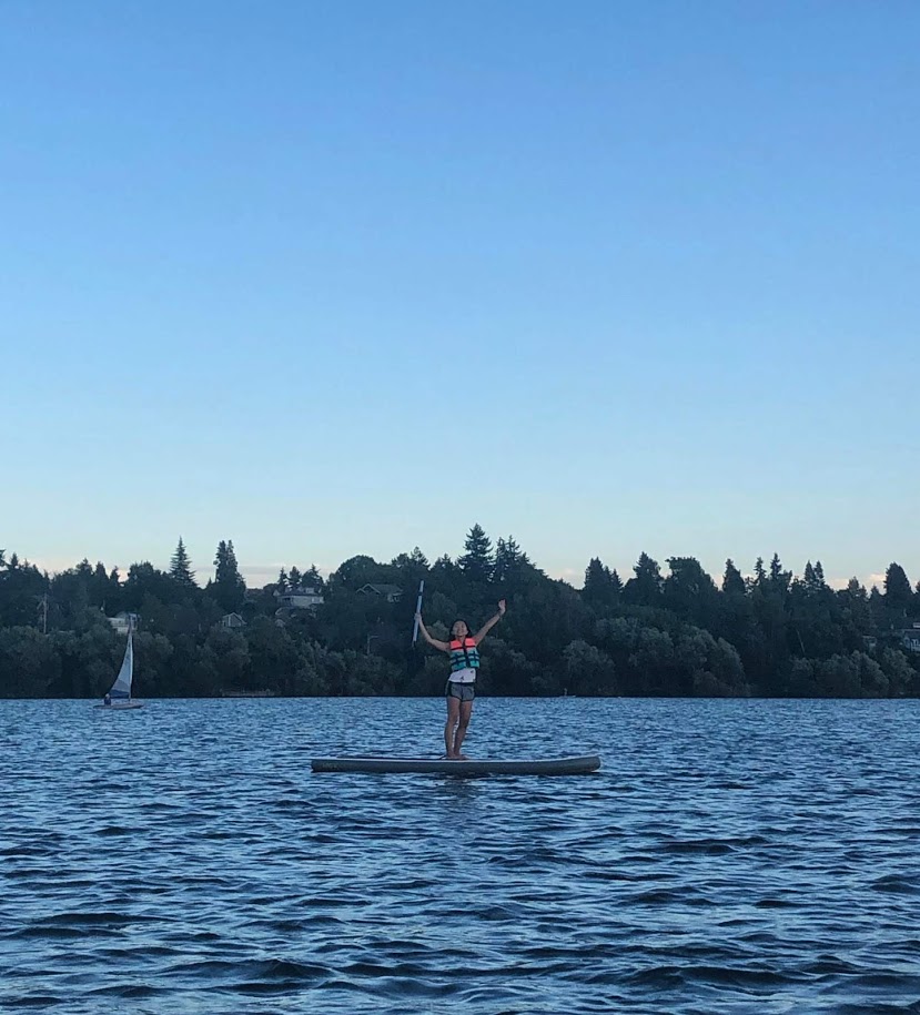Linda standing on paddle board in lake with both arms in the air